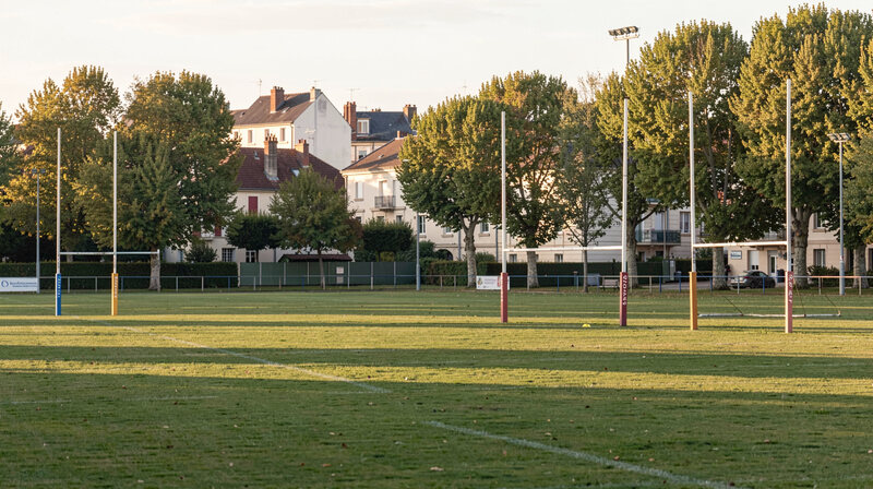 Vue large d'un terrain de rugby en fin de journée, poteaux de but dressés sur la pelouse, arbres et maisons du quartier en arrière-plan.